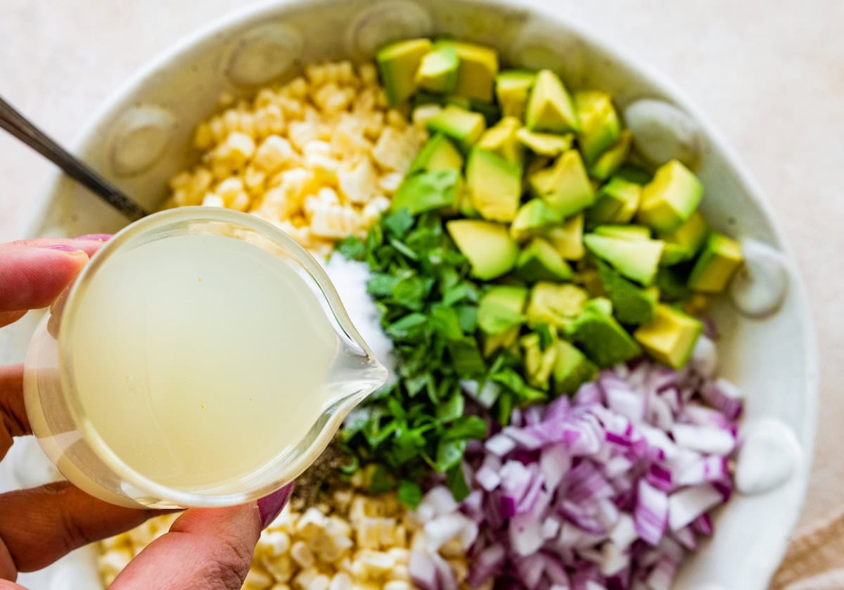 A woman's hand holds a small glass pitcher of lime juice over a large bowl of corn salad.