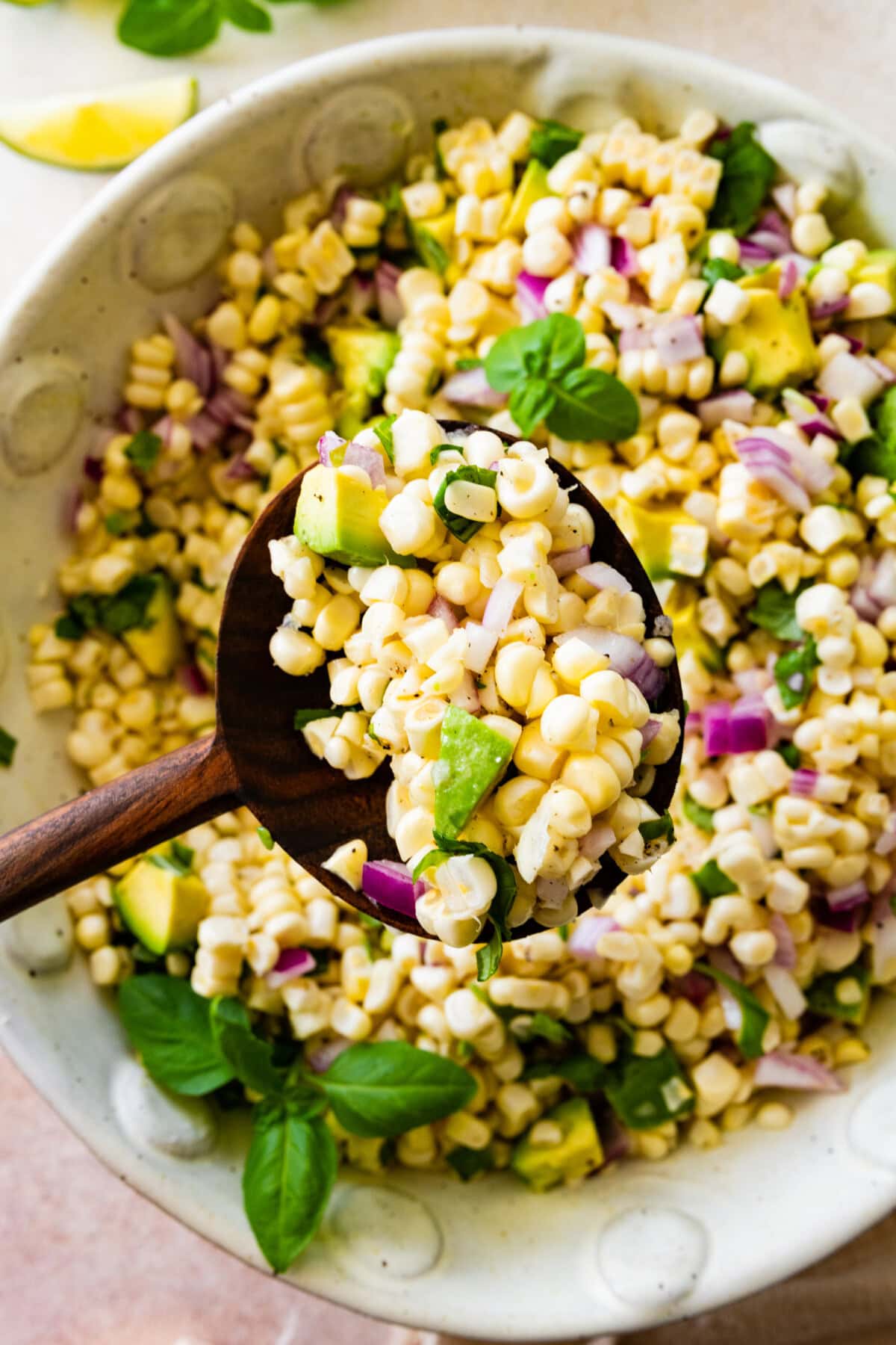 A large wooden spoon holding a portion of avocado corn salad over the bowl of the corn salad.