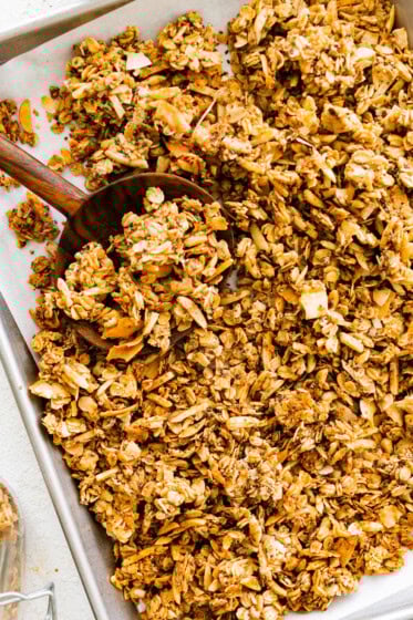 A baking sheet with a large wooden spoon taking a portion of coconut granola.