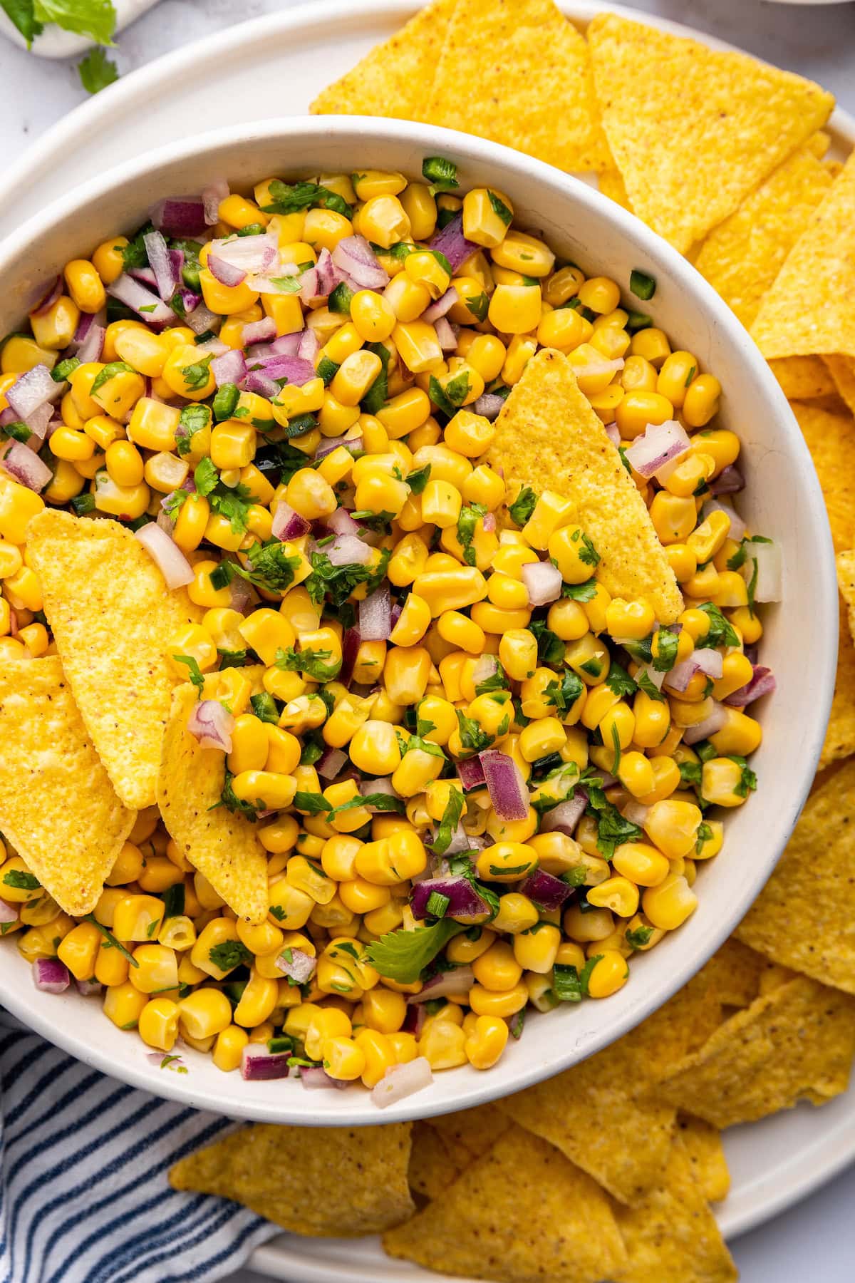 Corn mixed with red onion, cilantro and jalapeño in a white bowl served with tortilla chips.