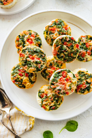 Multiple egg white bites topped with cheese and veggies rest on a white plate.