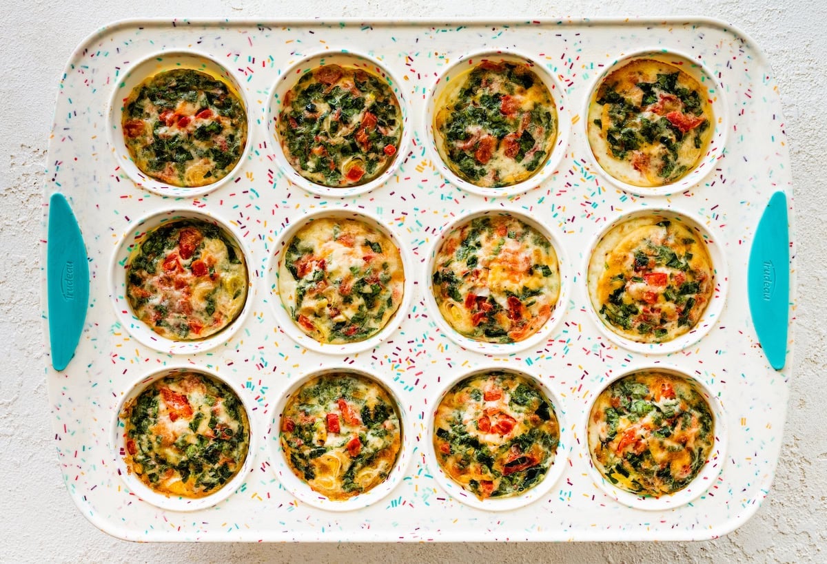 Twelve egg white bites in a muffin tray topped with cheese and vegetables.