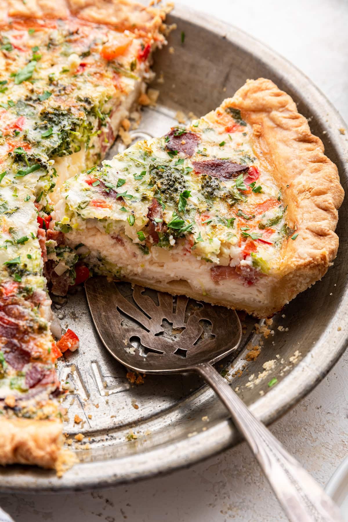 A slice of quiche being lifted from a metal pie dish using a silver pie server. The quiche has a golden-brown, flaky crust and is filled with a creamy mixture of egg whites, cottage cheese, and a variety of finely chopped vegetables, including broccoli, red pepper, and white onion. It also contains shredded mozzarella cheese, chopped cooked turkey bacon, and fresh herbs like parsley.