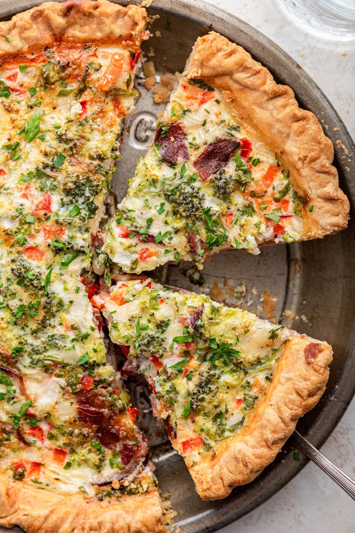 A quiche in a metal pie dish, with a slice partially removed. The quiche has a golden-brown, flaky crust and topped with fresh herbs.