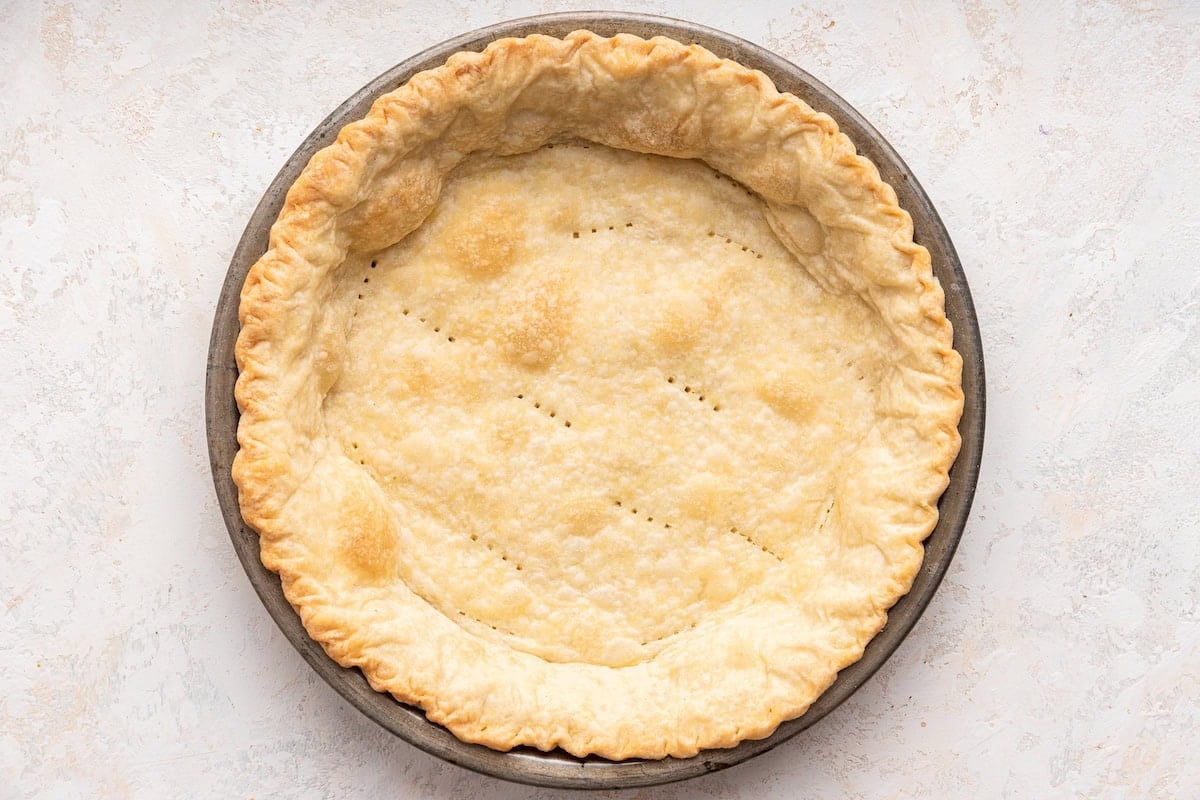 A pre-baked quiche crust in a metal pie dish. The crust is golden-brown and flaky, with small fork pricks evenly distributed across the bottom to prevent it from puffing up during baking. The edges of the crust are crimped, giving it a decorative, textured border. The crust is empty and ready to be filled with the quiche mixture.
