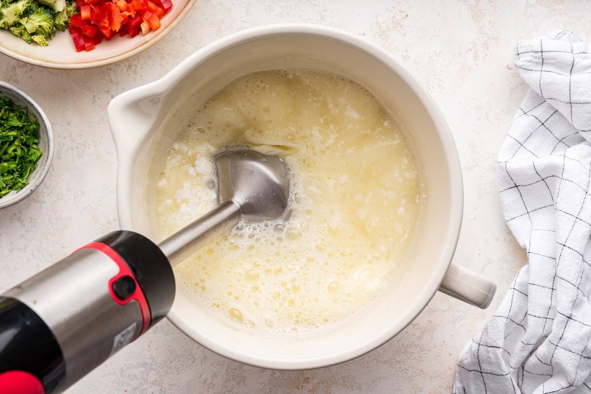 A white mixing bowl contains a frothy, pale yellow mixture of egg whites and cottage cheese being blended with an immersion blender.
