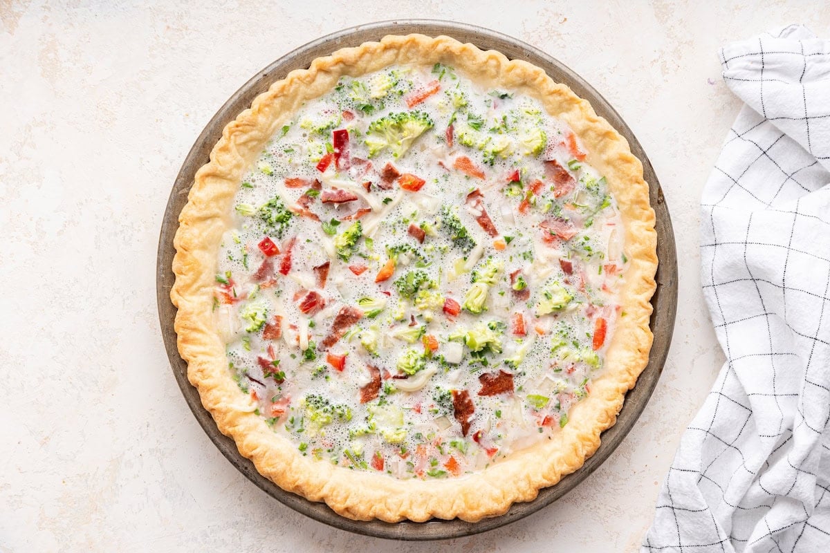 A pre-baked quiche ready to go into the oven. The quiche crust is golden-brown and flaky, filled with a creamy mixture of egg whites, cottage cheese, finely chopped broccoli, red pepper, white onion, shredded mozzarella cheese, and chopped cooked turkey bacon.