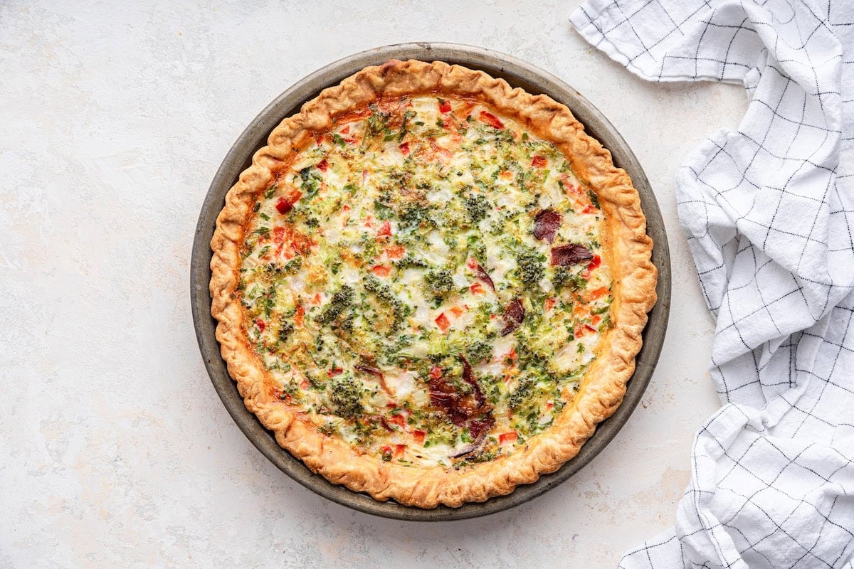 A freshly baked quiche in a metal pie dish. The quiche has a golden-brown, flaky crust and a well-set filling. The filling consists of egg whites, cottage cheese, finely chopped broccoli, red pepper, white onion, shredded mozzarella cheese, and chopped cooked turkey bacon. The top is slightly browned, with visible bits of vegetables and herbs.