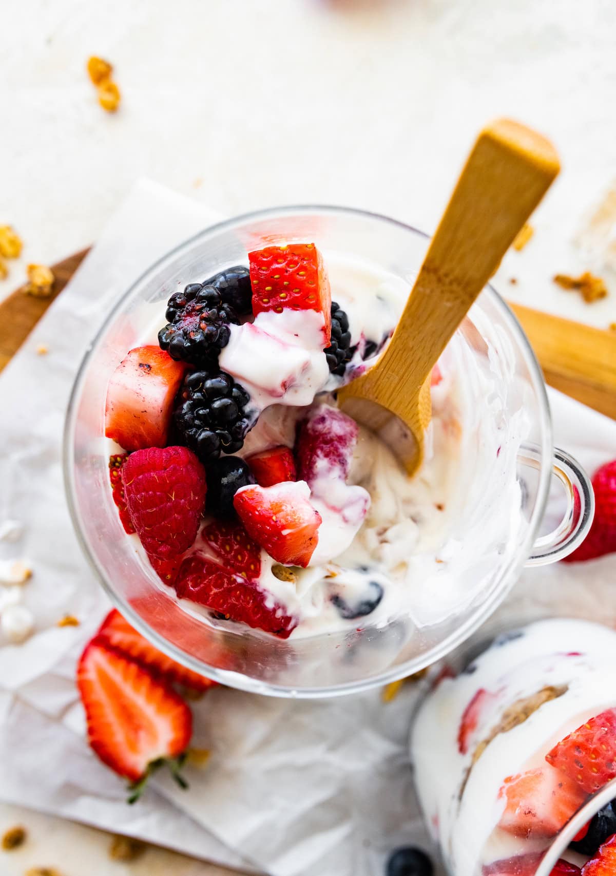 A glass cup of a half-eaten fruit and yogurt parfait with fresh berries and a wooden spoon.