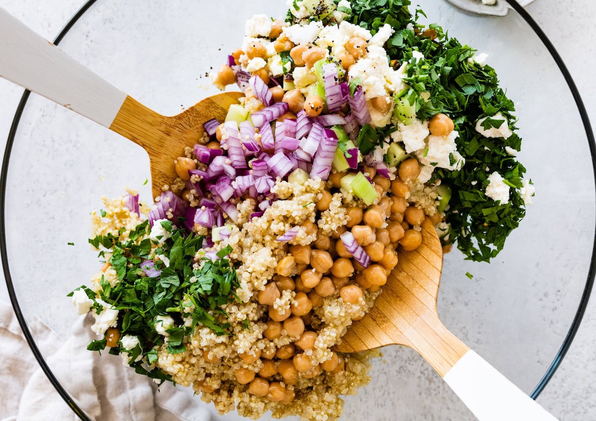 Two wooden serving spoons in a large glass bowl of salad ingredients that include quinoa, chickpeas, and fresh vegetables.