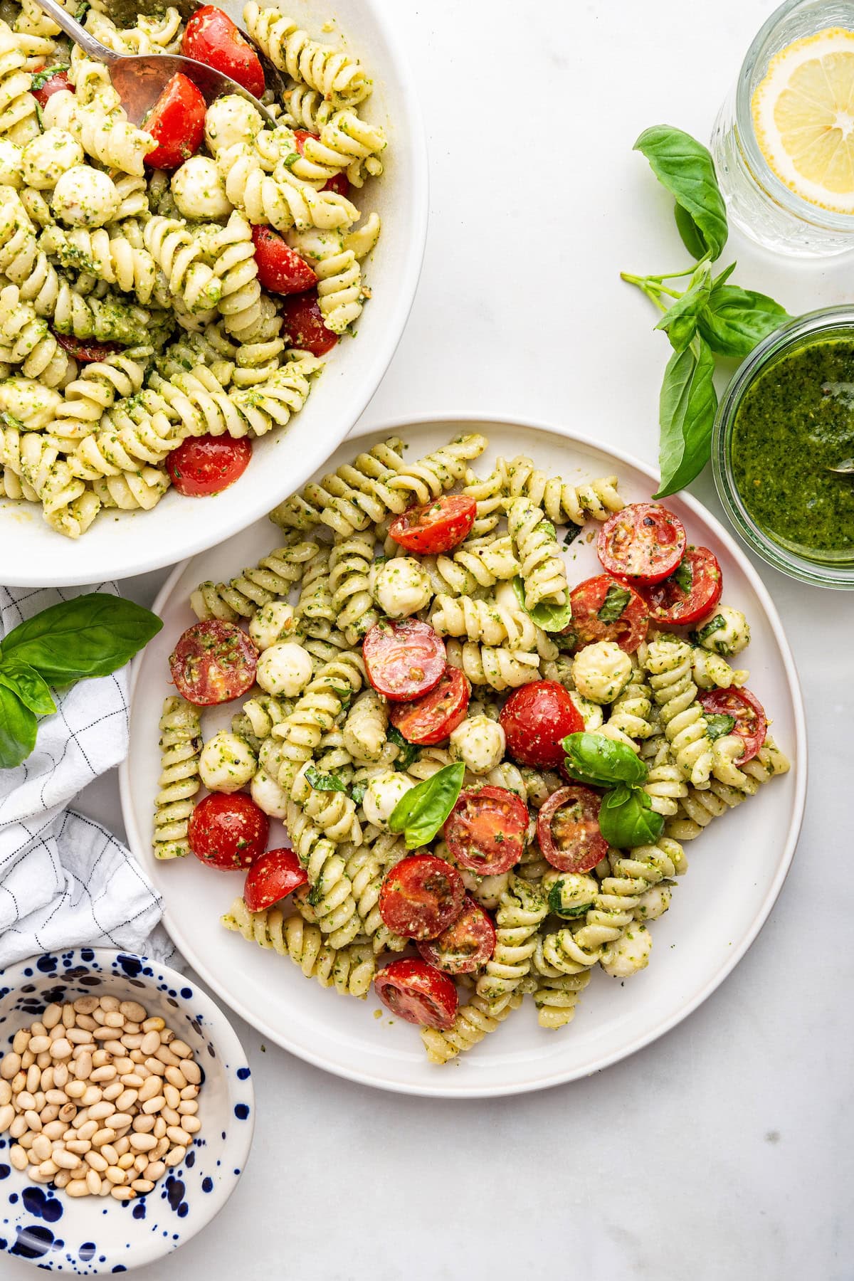 A serving of pesto pasta salad on a white plate near a bowl of the pasta salad.