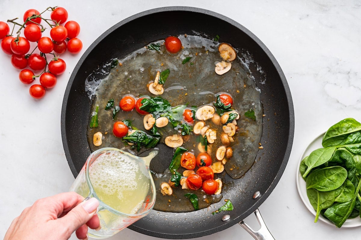 Egg whites being poured into a skillet with mushrooms, spinach, and cherry tomatoes.