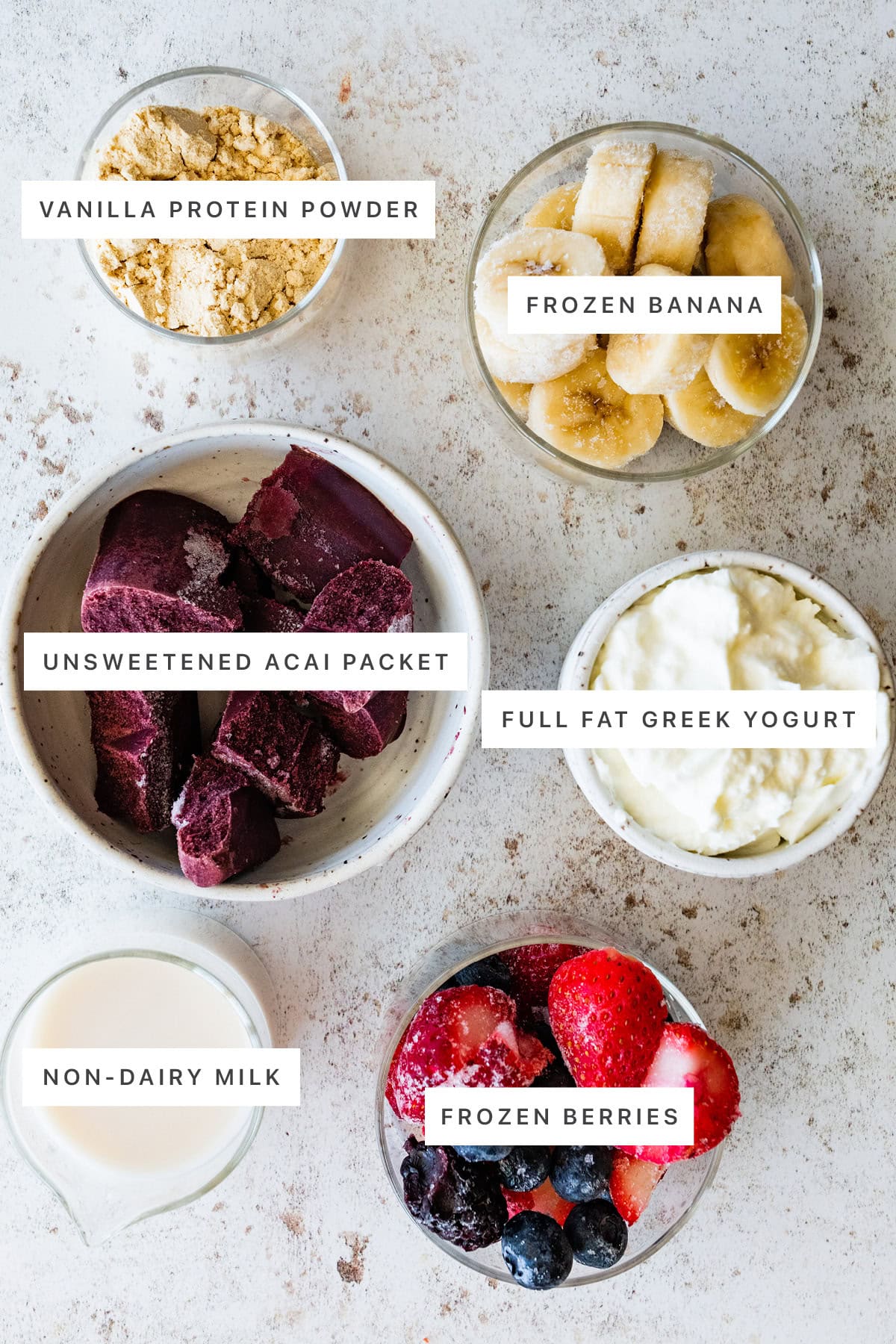 Ingredients measured out to make Protein Acai Bowl: vanilla protein powder, frozen banana, unsweetened acai packet, full fat greek yogurt, non-dairy milk and frozen berries.