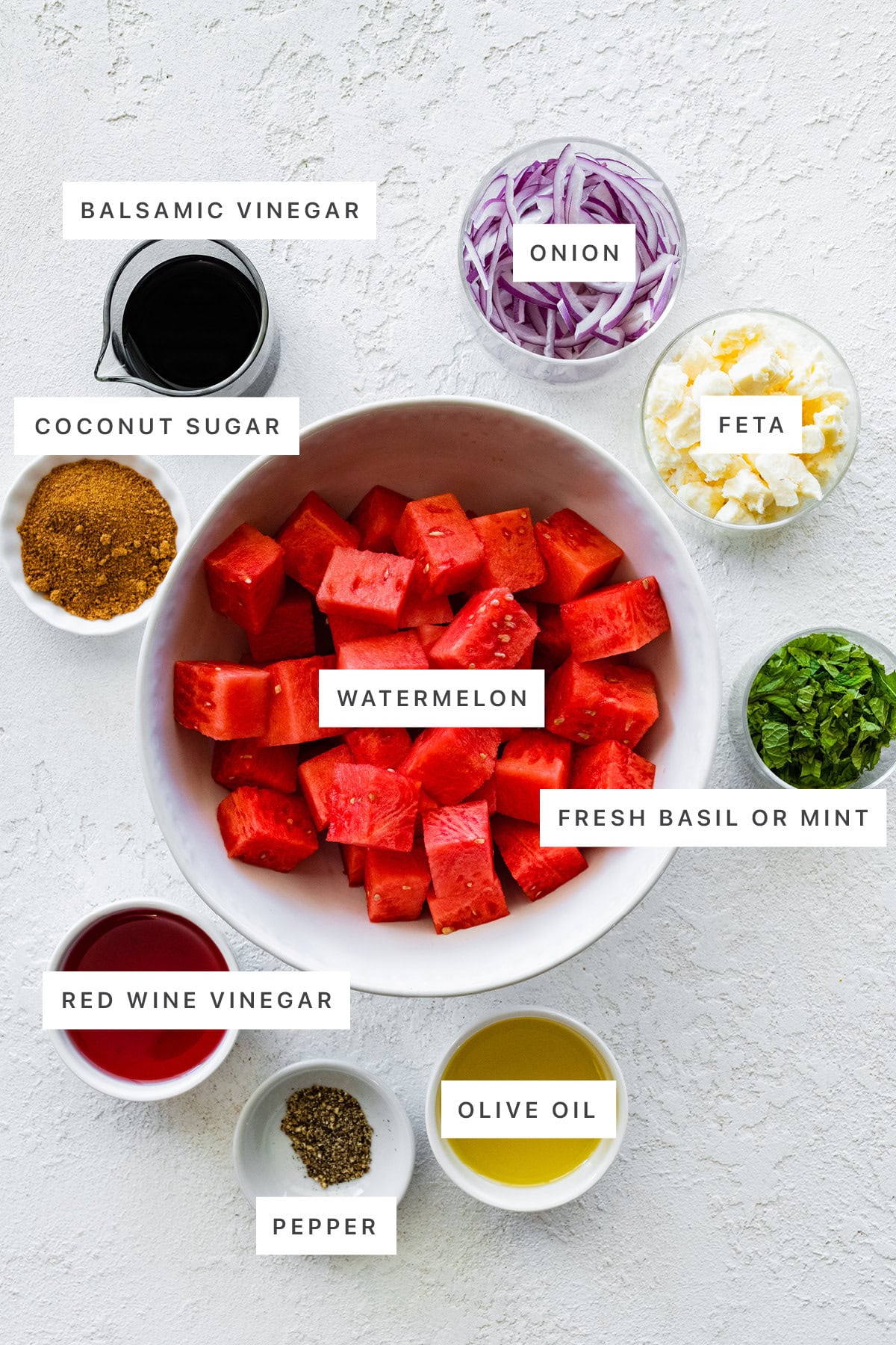 Ingredients measured out to make Watermelon Salad with Feta and Basil: balsamic vinegar, coconut sugar, onion, feta, watermelon, fresh mint or basil, red wine vinegar, pepper and olive oil.