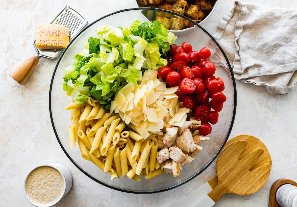 Pasta noodles, cheese, lettuce, cherry tomatoes, and chicken in a large glass bowl.