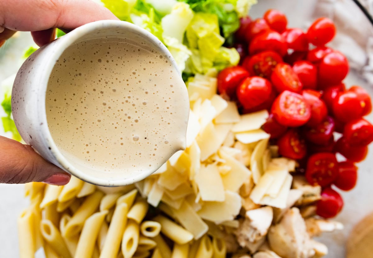 A womans hand pours a caesar dressing over a large bowl of chicken caesar pasta salad.