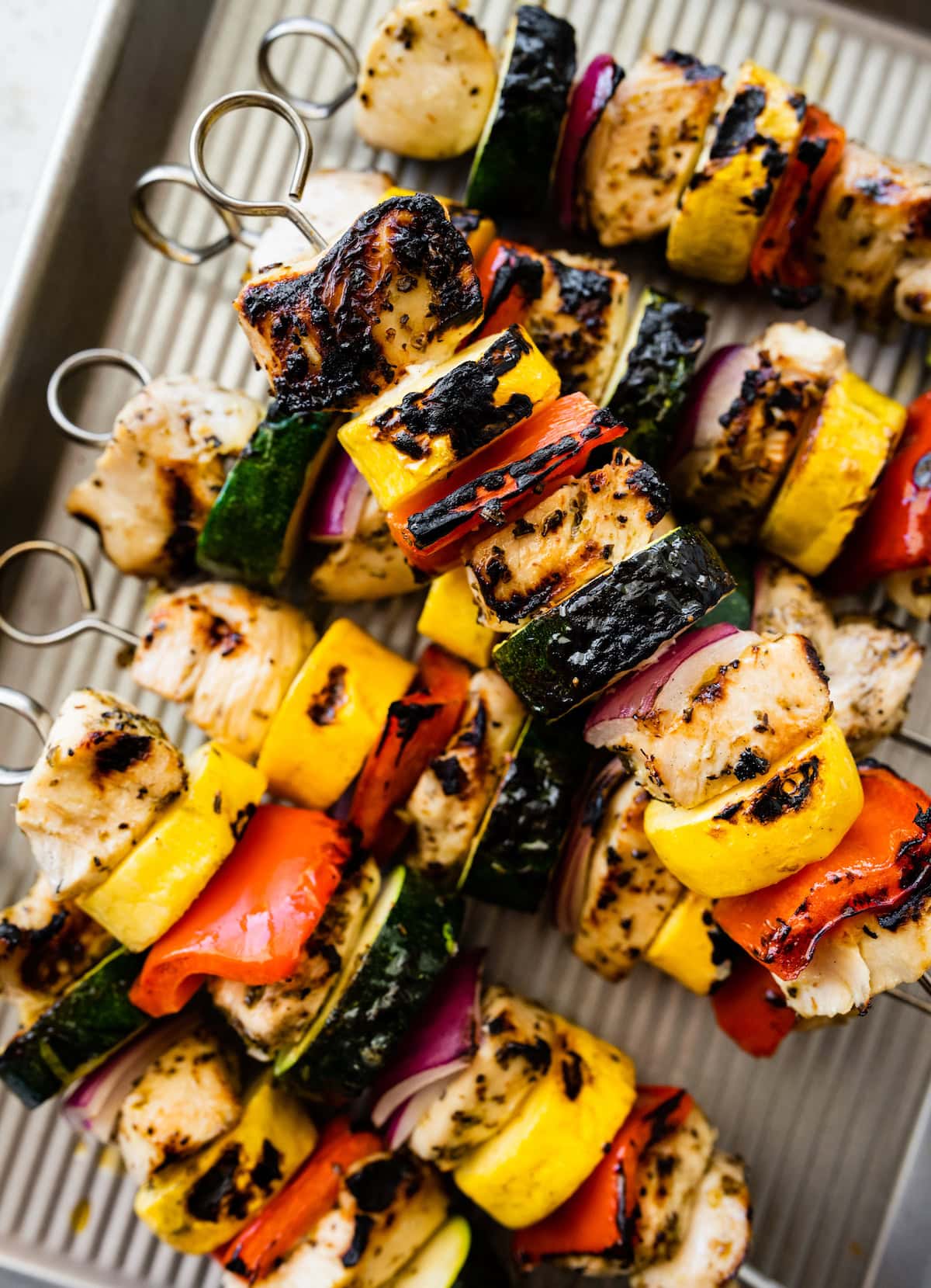 Six grilled chicken kabobs on a baking sheet. The kabobs combine chicken, yellow squash, zucchini, red pepper and onion.