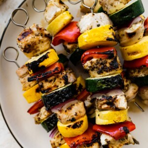 Six grilled chicken kabobs on a white plate. The kabobs combine chicken, yellow squash, zucchini, red pepper and onion.