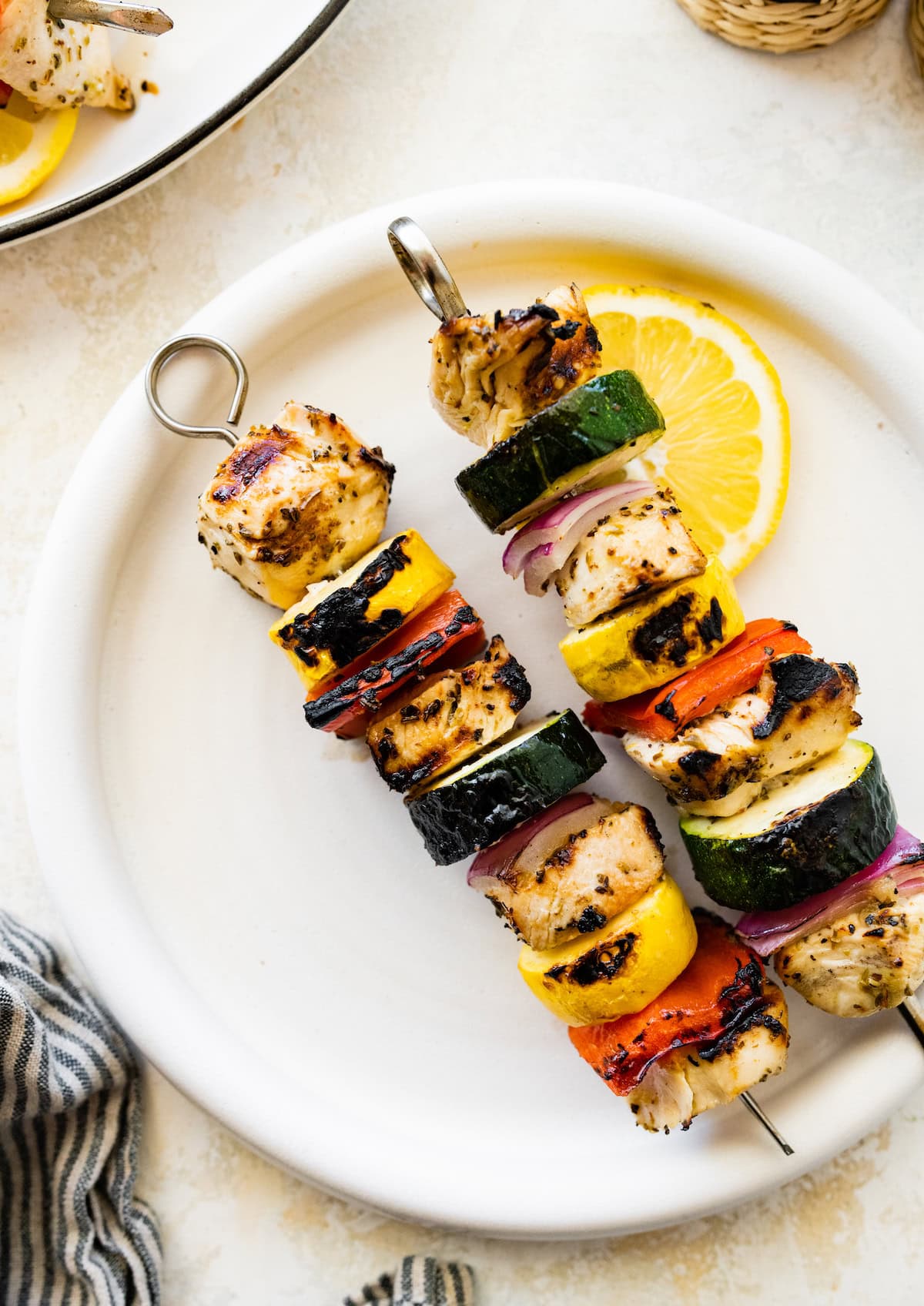 Two grilled chicken kabobs on a white plate served with a lemon slice. The kabobs combine chicken, yellow squash, zucchini, red pepper and onion.