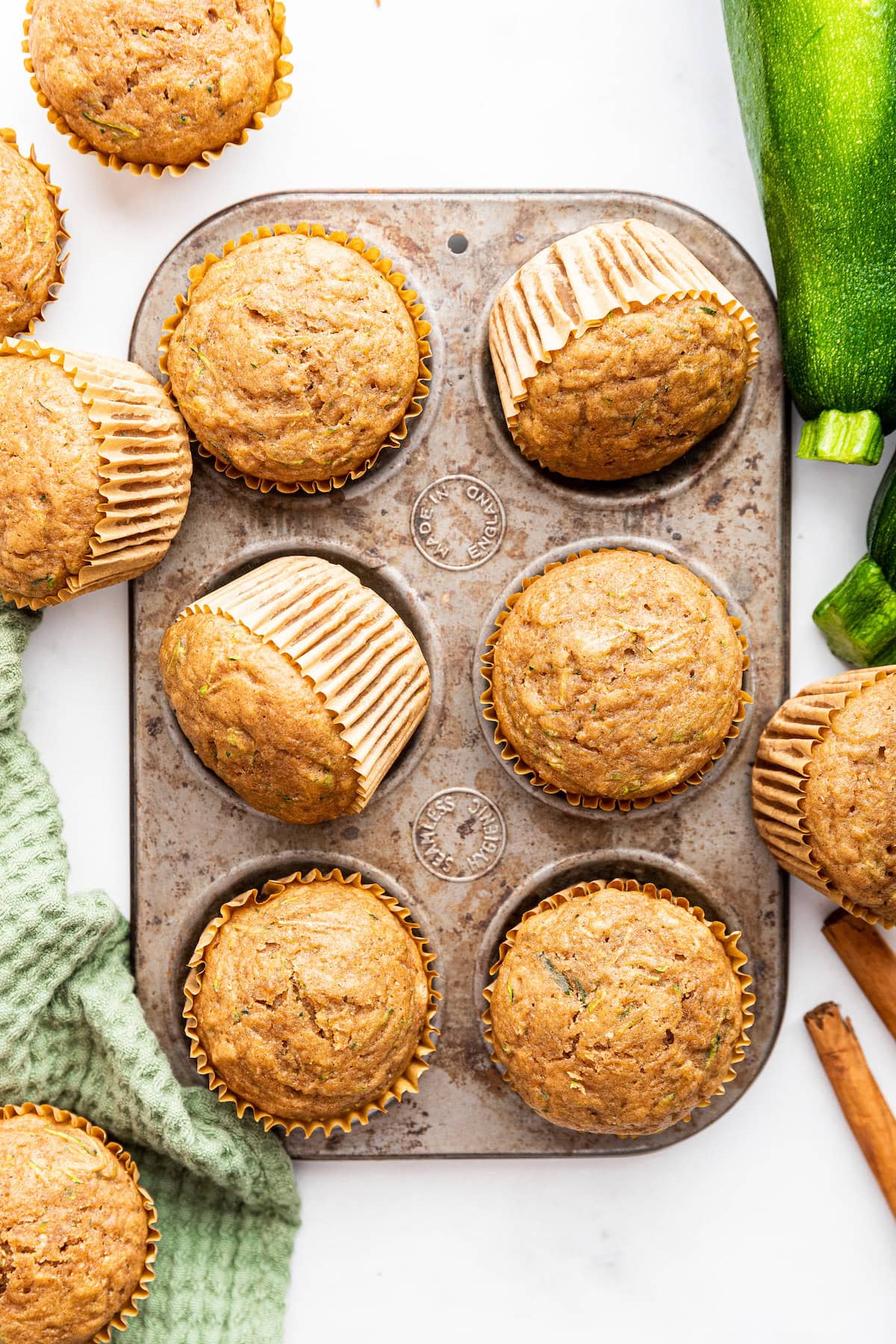 Six zucchini muffins in a muffin tin with more muffins and fresh zucchini nearby.