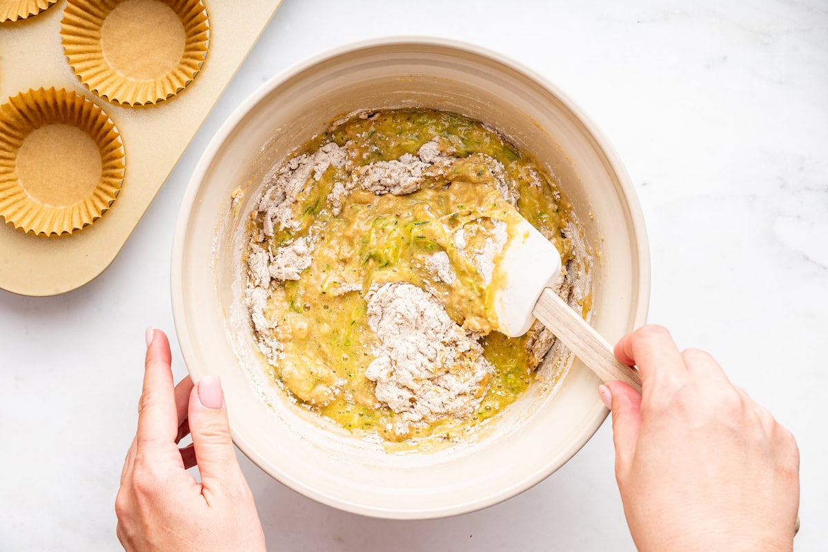 A womans hand uses a silicone spatula to mix flour, zucchini, and egg in a large mixing bowl.