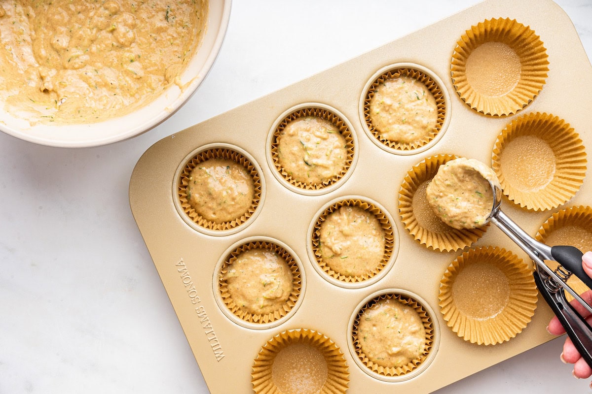 A womans hand uses a cookie scoop to portion zucchini muffin batter into a muffin tin.