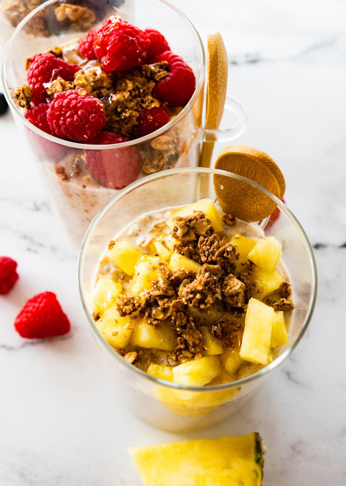 Two glass cups of kefir overnight oats. One cup is topped with fresh raspberries and granola while the other cup has fresh pineapple.