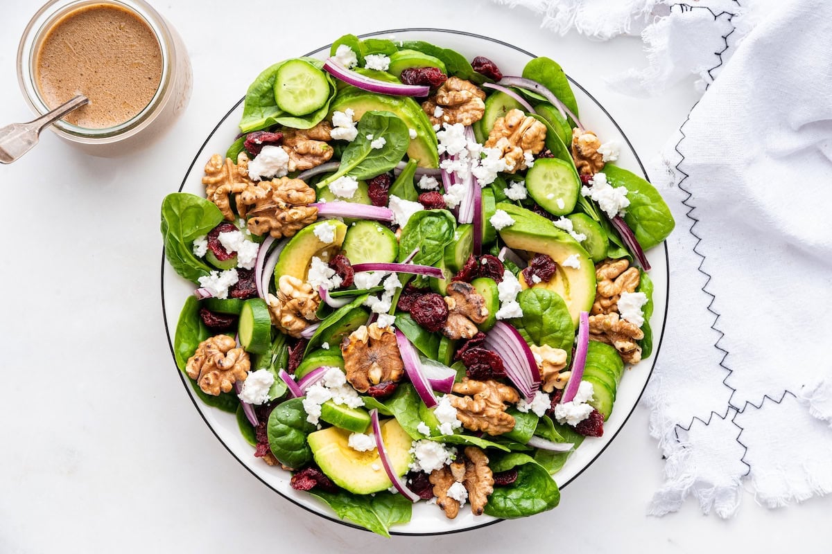 A bowl of spinach salad with ingredients like red onion, walnut, avocado, and cranberries.