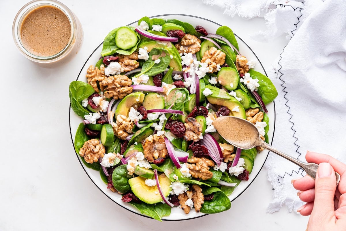 A womans hand uses a spoon to drizzle dressing over a spinach salad.