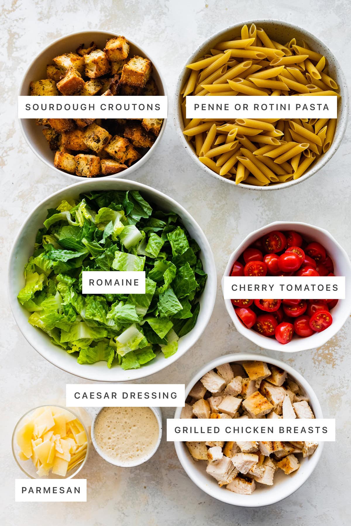 Ingredients measured out to make Chicken Caesar Pasta Salad: sourdough croutons, penne pasta, romaine, cherry tomatoes, parmesan, caesar dressing and grilled chicken breasts.