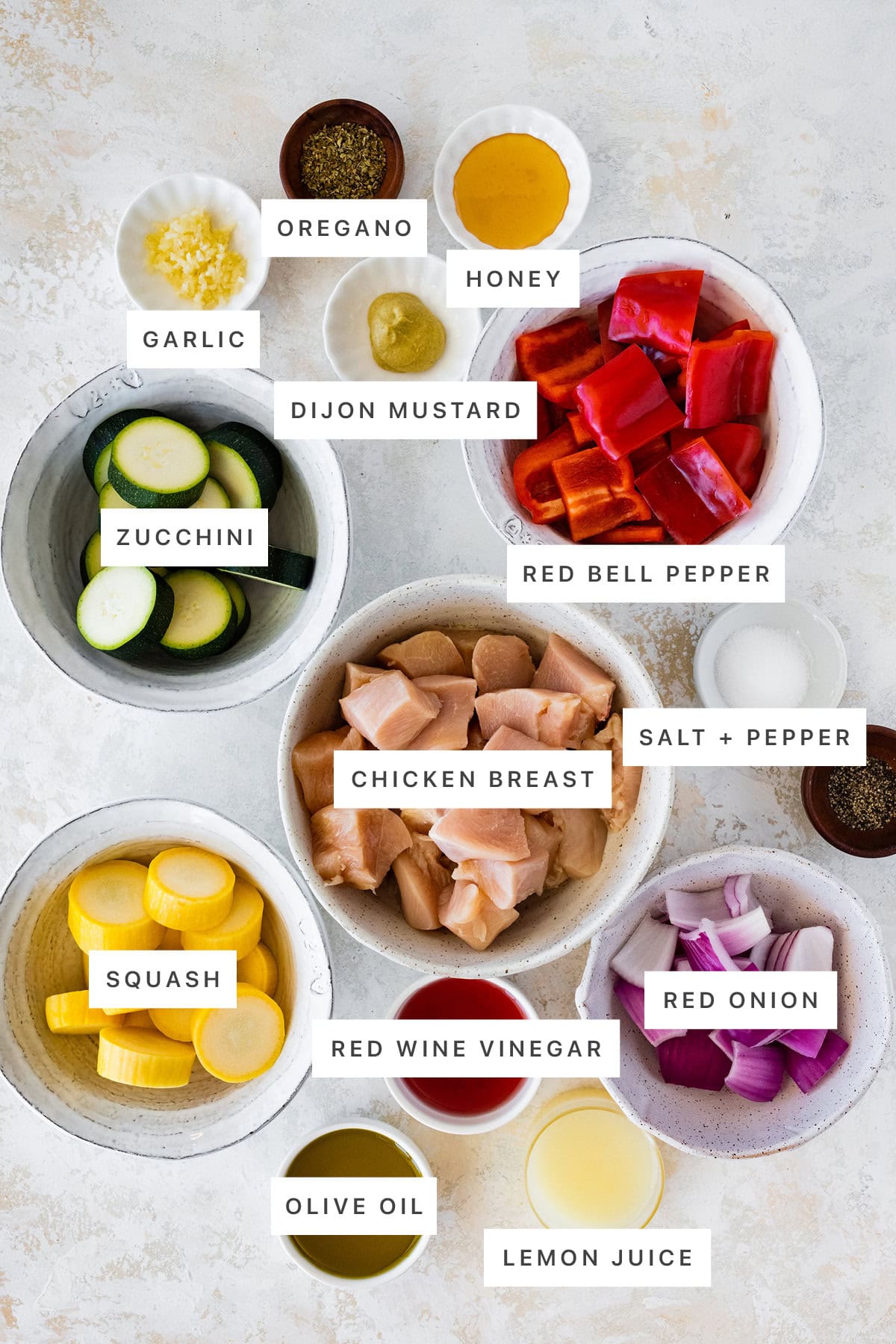 Ingredients measured out to make Grilled Chicken Kabobs: garlic, oregano, honey, dijon, bell pepper, zucchini, chicken breast, salt, pepper, squash, red wine vinegar, red onion, olive oil and lemon juice.