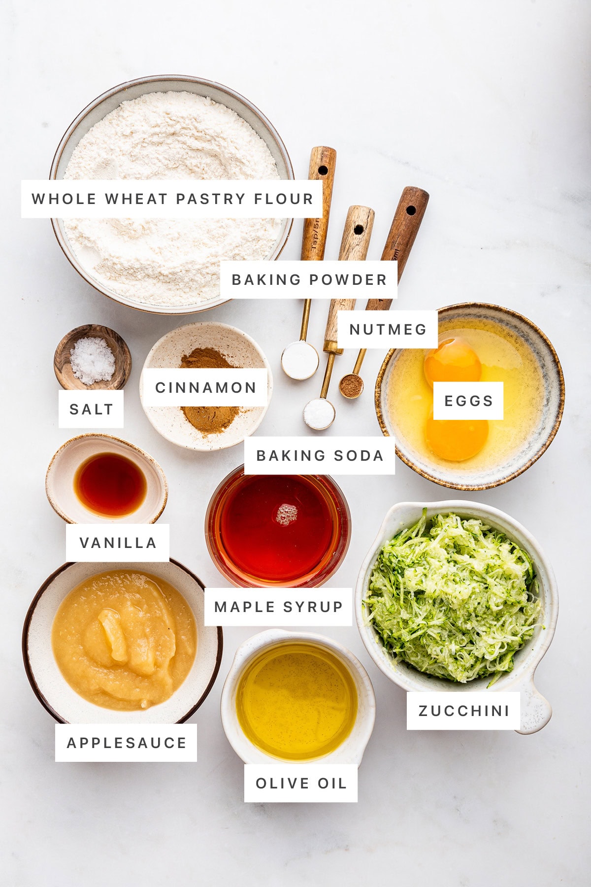 Ingredients measured out to make Healthy Zucchini Muffins: whole wheat pastry flour, baking powder, nutmeg, cinnamon, baking soda, salt, eggs, vanilla, maple syrup, zucchini, applesauce and olive oil.