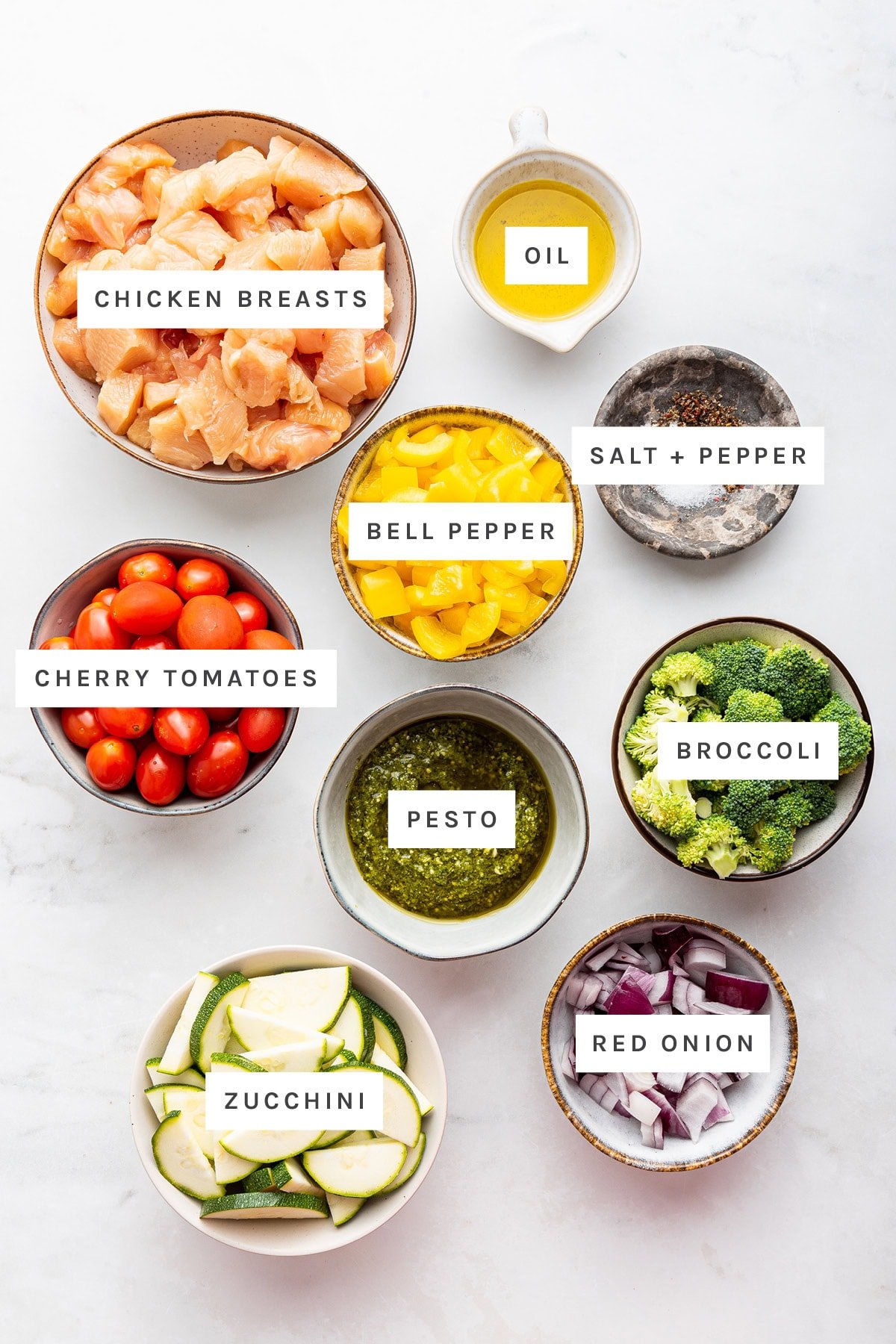 Ingredients measured out to make Pesto Chicken: chicken breasts, oil, bell pepper, salt, pepper, cherry tomatoes, pesto, broccoli, zucchini and red onion.