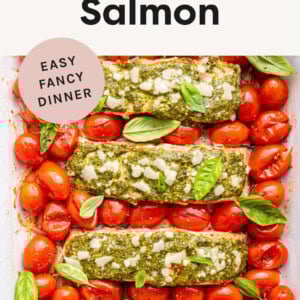 Four pieces of salmon, topped with pesto, cheese and surrounded by tomatoes in a roasting pan.
