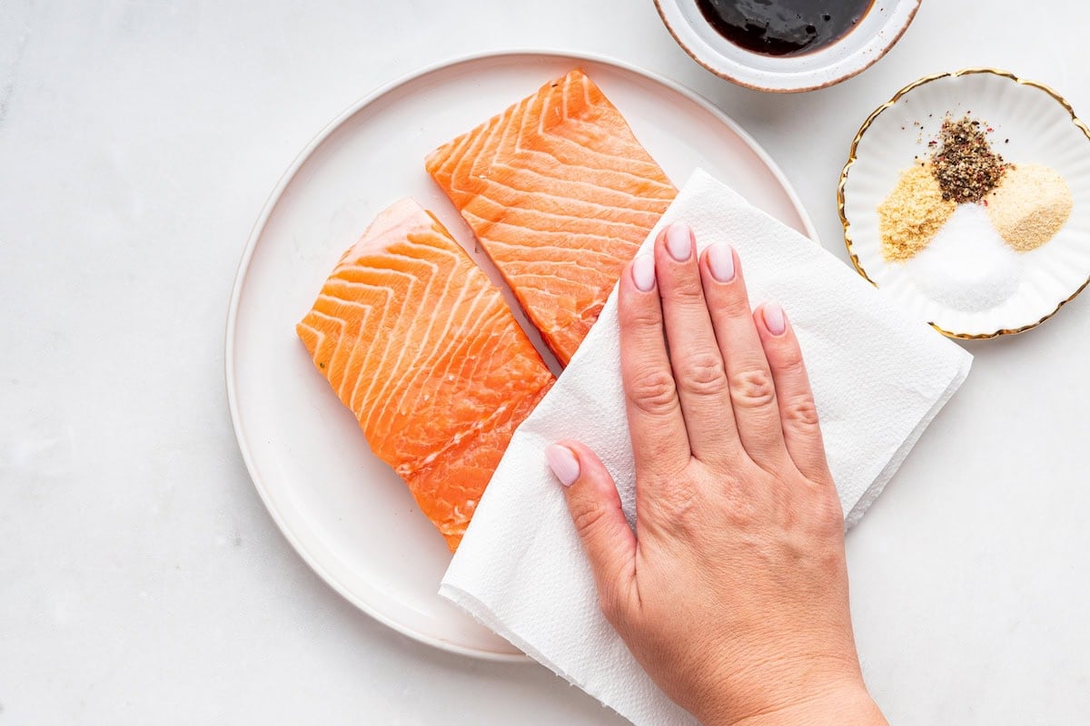 A woman's hand uses a paper towel to pat dry two raw salmon filets.