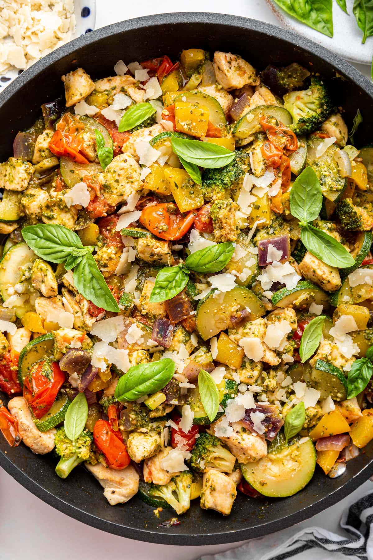 A large skillet of cooked pesto chicken with multiple vegetables and topped with fresh basil leaves.