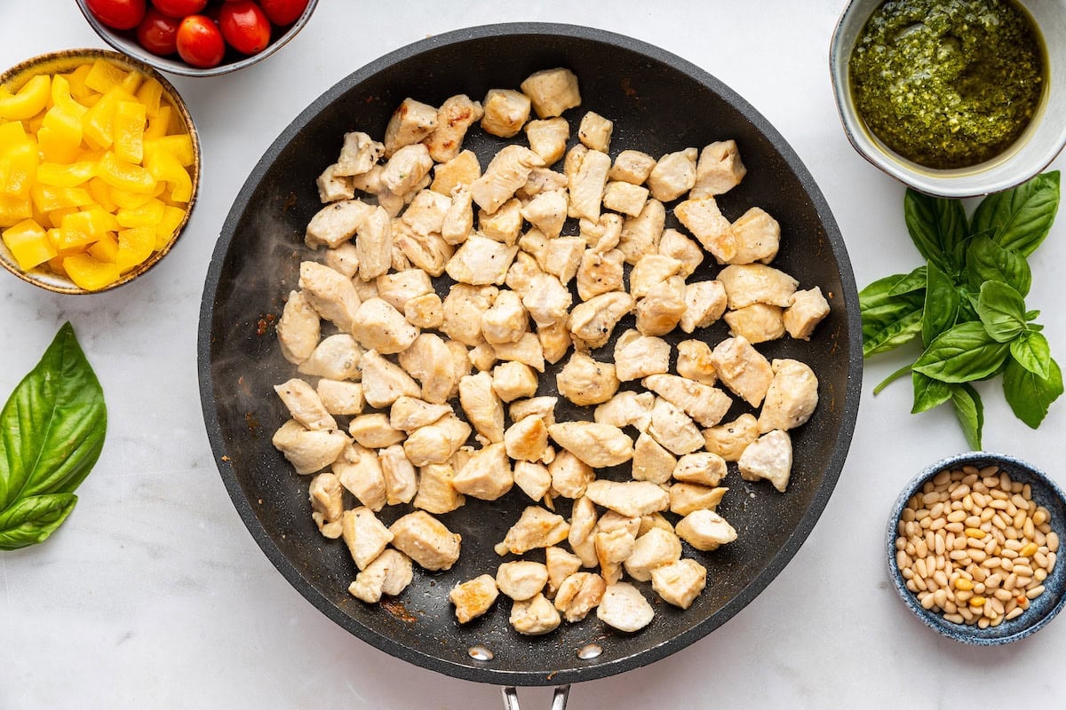 A large skillet of cooked chicken near small bowls of pine nuts, pesto, bell peppers, and tomato.