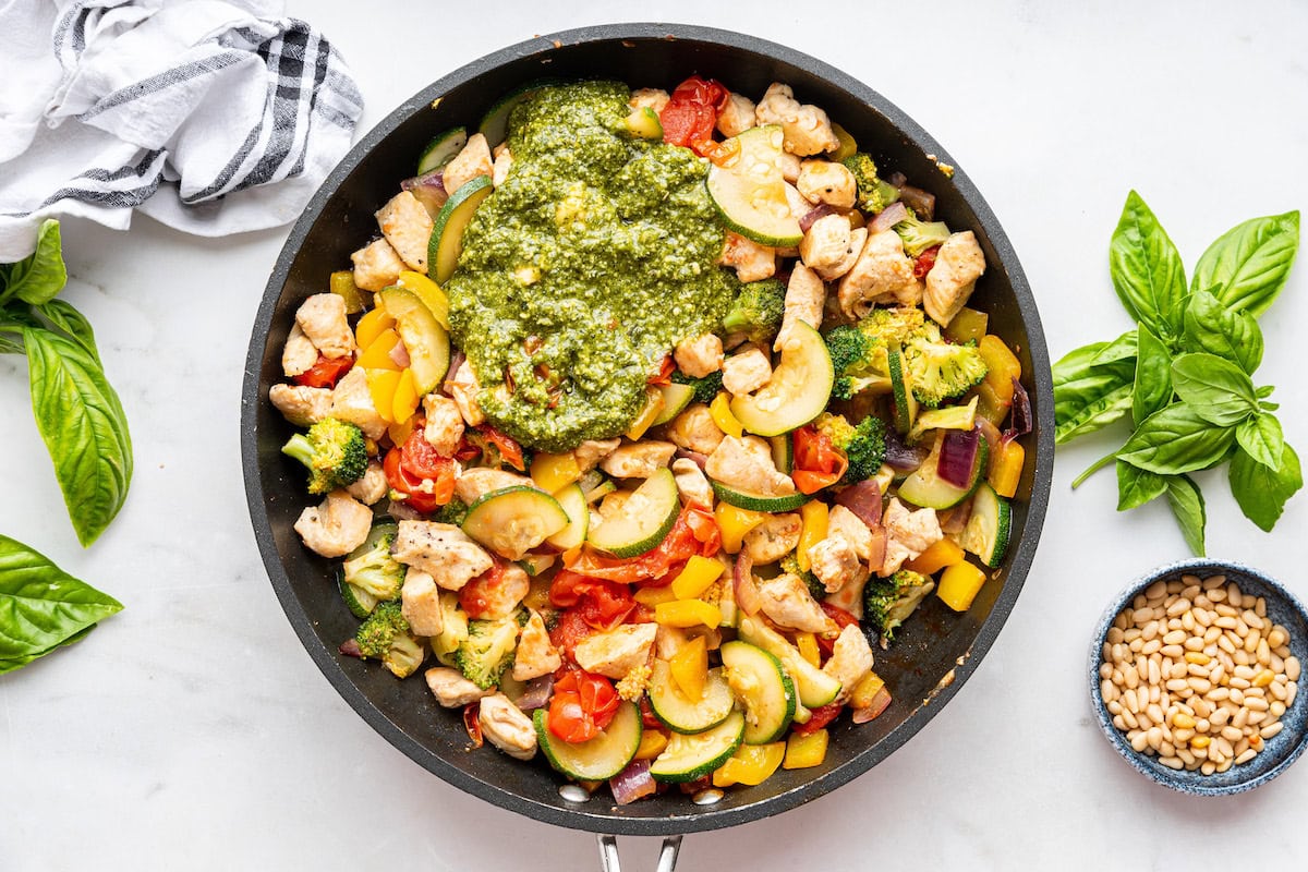 A large skillet of cooked pesto chicken with multiple vegetables and topped with fresh basil leaves.