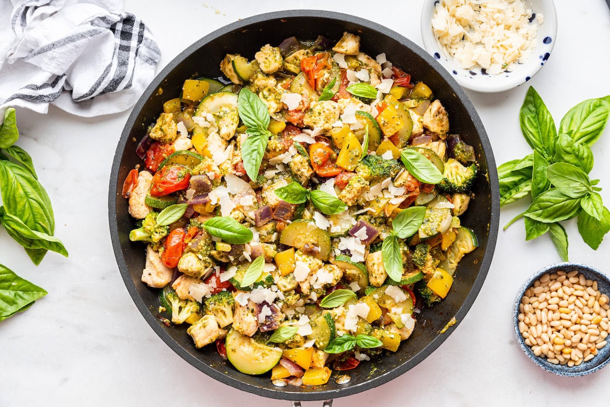 A large skillet of cooked pesto chicken with multiple vegetables and topped with fresh basil leaves.