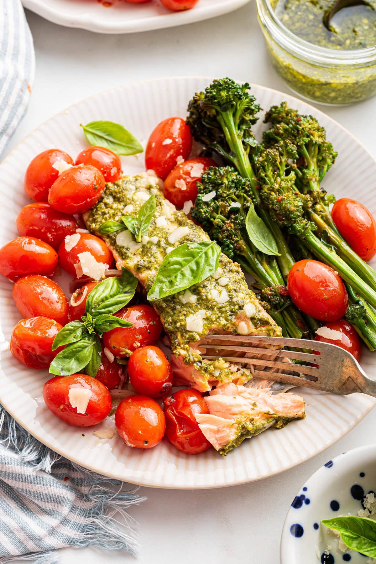 A plate of salmon covered in pesto with a side of cherry tomatoes and broccolini. There's a fork taking a portion of the salmon.