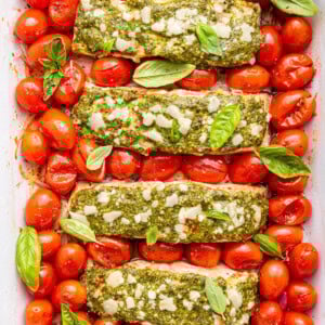Four salmon filets covered in pesto and surrounded by cherry tomatoes in a baking dish.