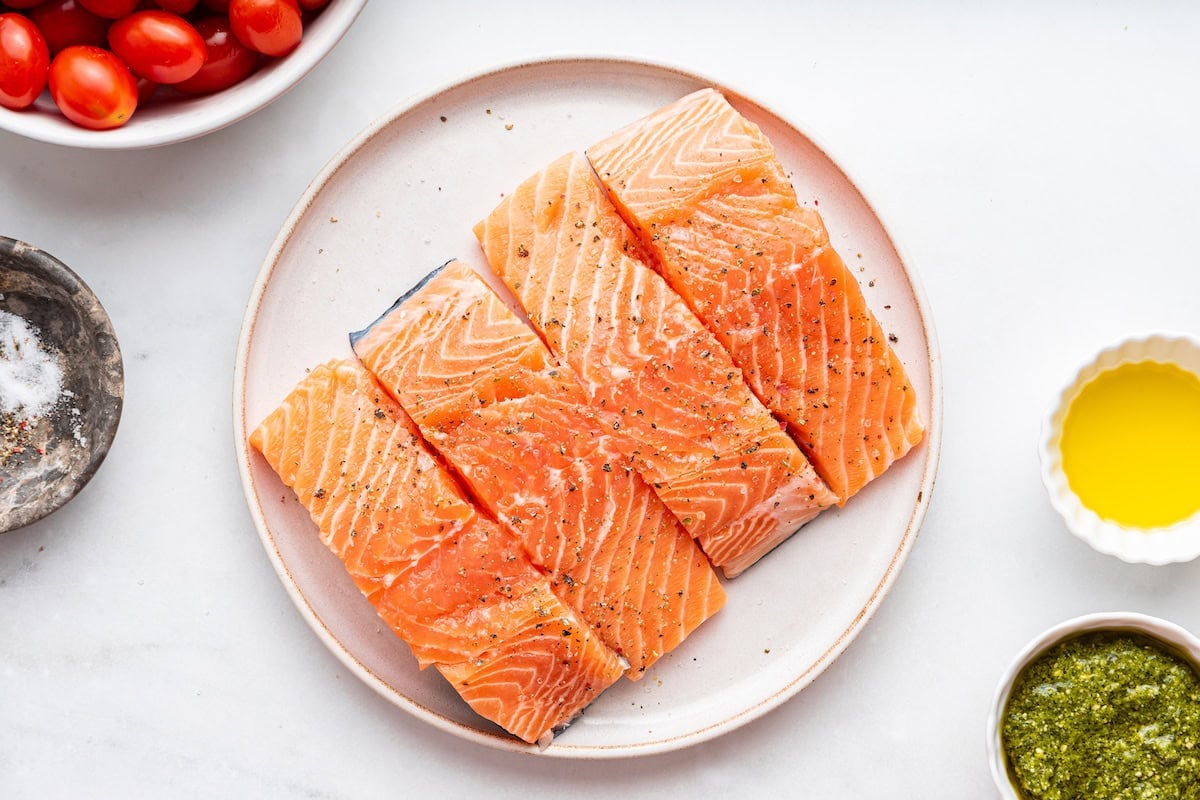 Four raw salmon filets with seasonings on a plate.