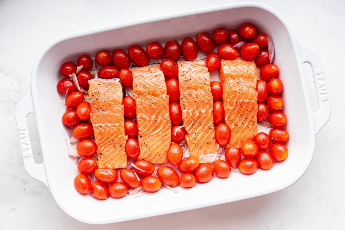 Four raw salmon filets in a large baking dish with cherry tomatoes and shallots.