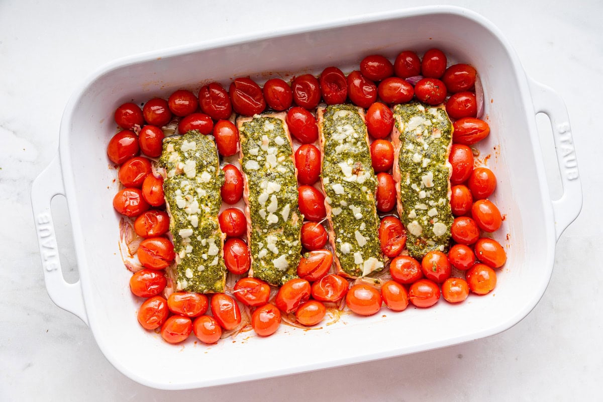 Four raw salmon filets covered in pesto in a large baking dish with cherry tomatoes and shallots.