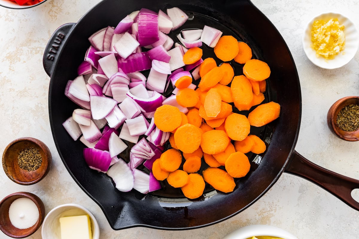 A cast iron skillet with uncooked sliced carrots, and red onion.