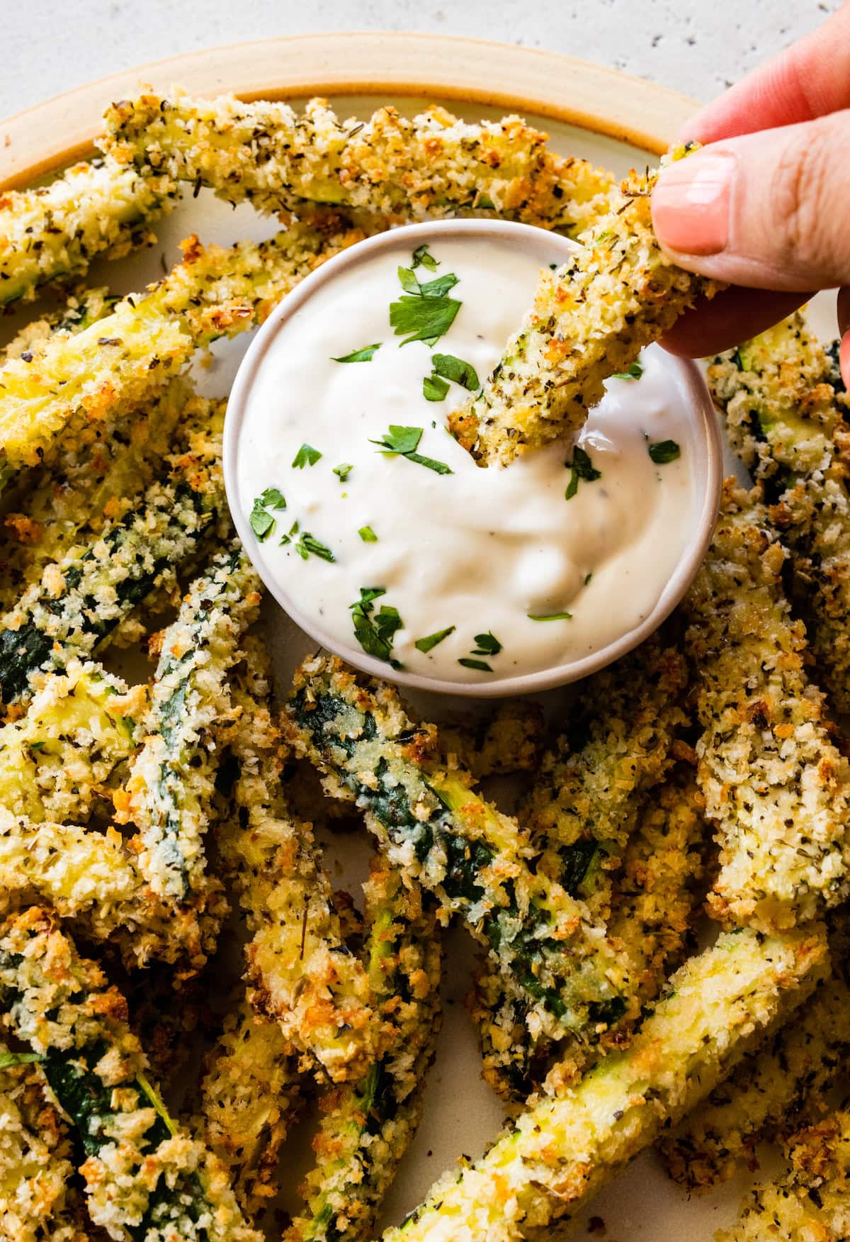 A woman's hand dips a zucchini fry in a ranch dip on a plate full of zucchini fries.