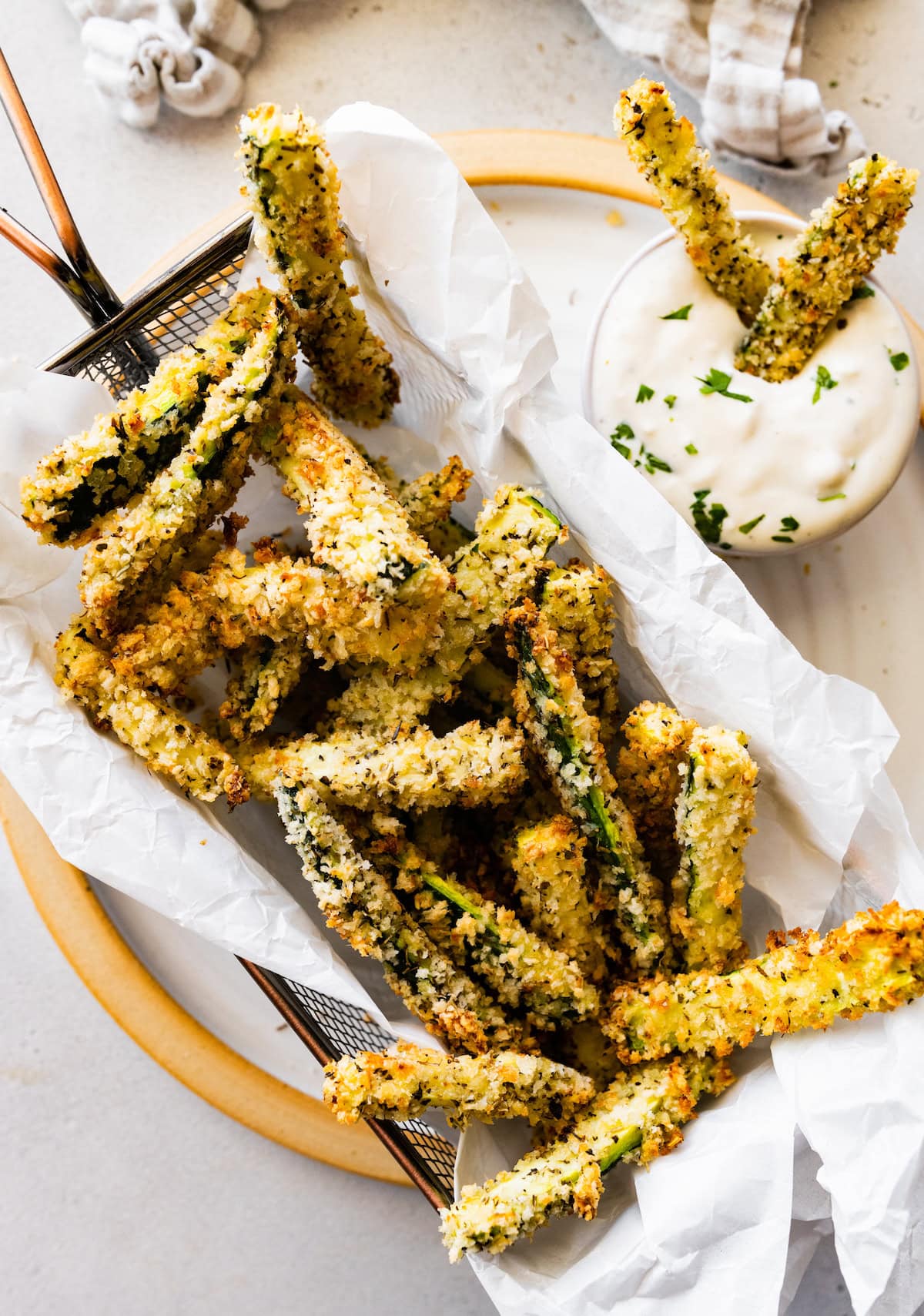 A metal frying basket on a plate full of crispy zucchini fries near a dipping sauce.