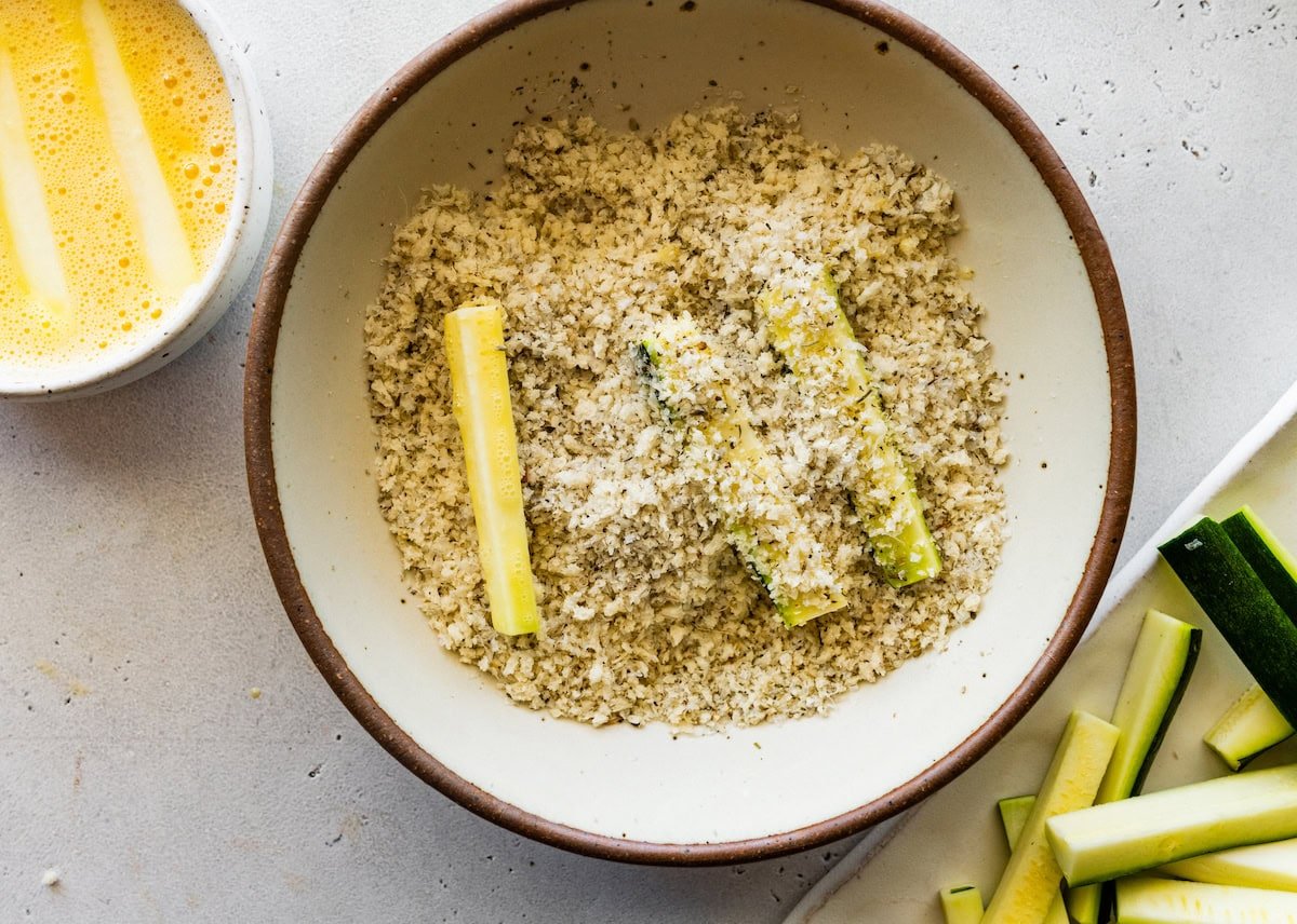 Zucchini fries in a large mixing bowl of bread crumbs, parmesan cheese, and seasoning.