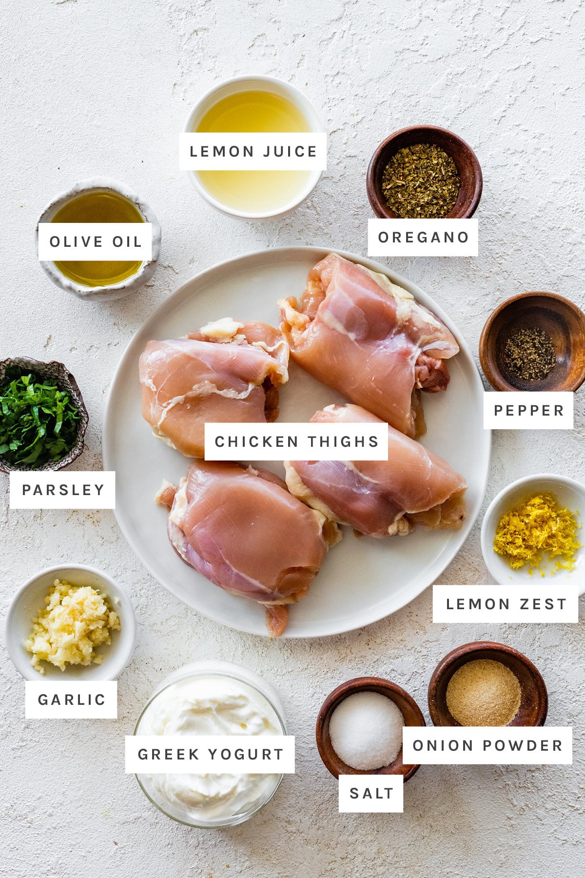 Ingredients measured out to make Yogurt Marinated Chicken: lemon juice, olive oil, oregano, parsley, chicken thighs, pepper, lemon zest, garlic, Greek yogurt, salt and onion powder.