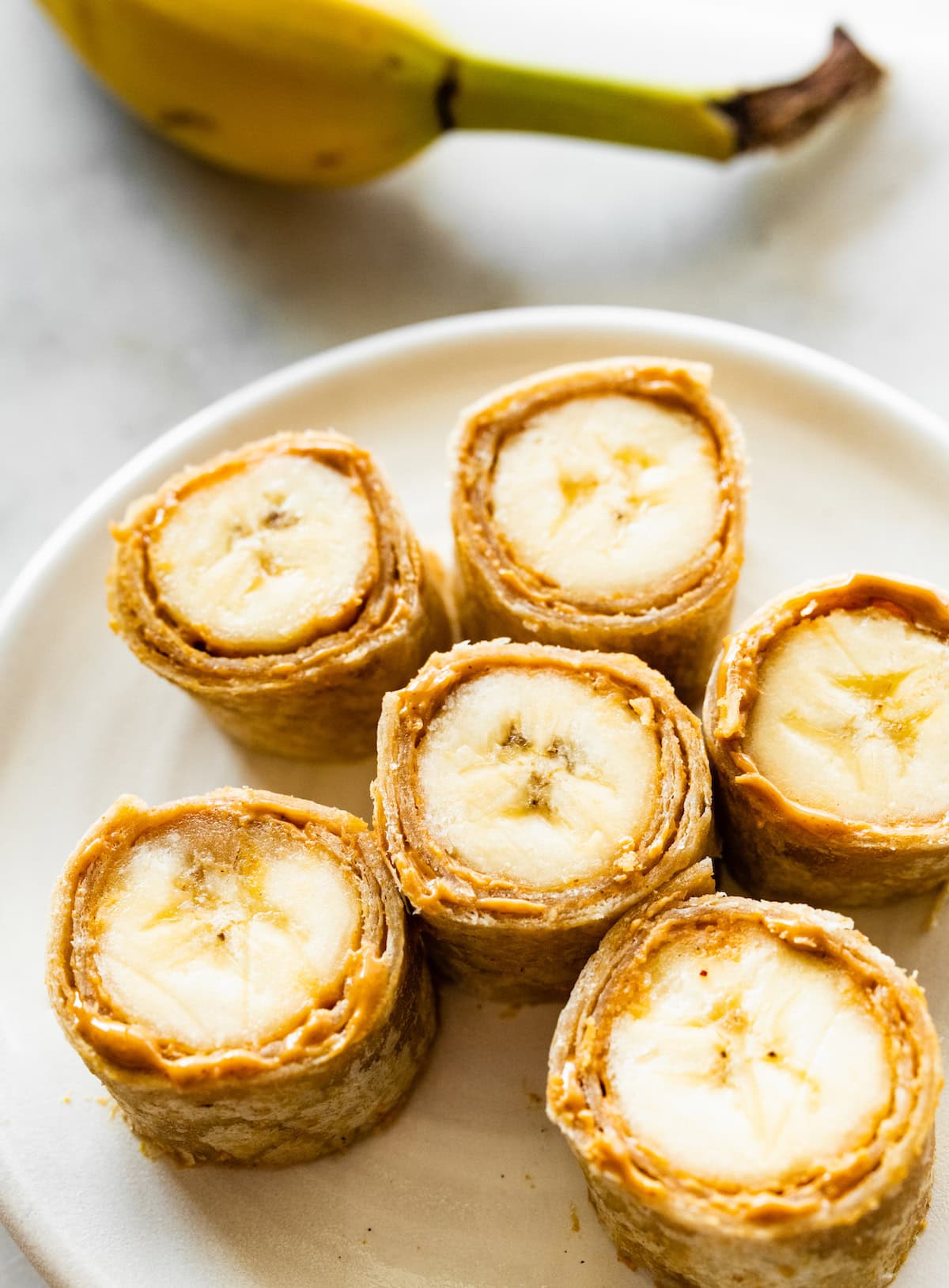 Six banana sushi roll-ups on a plate. The bananas are wrapped in a tortilla with a nut butter inside.